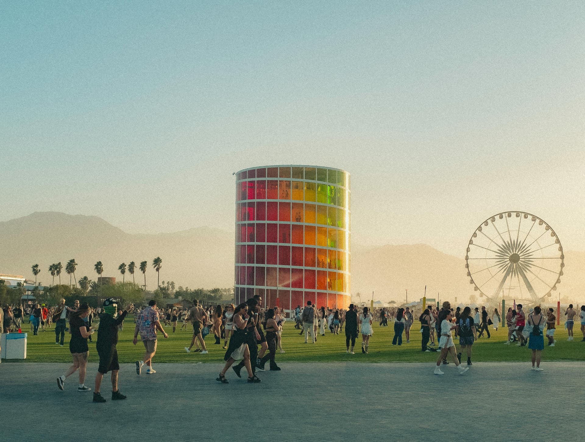 Coachella's rainbow-paneled Spectra tower rises above festival crowds at golden hour, with the Ferris wheel visible in the distance