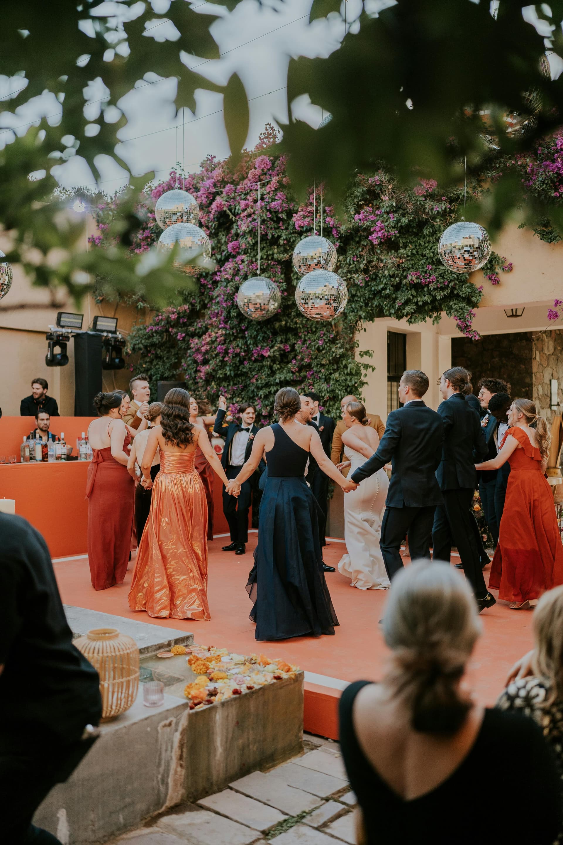 Wedding party holding hands in a circle on a coral dance floor, surrounded by hanging disco balls and bougainvillea in bloom at golden hour.