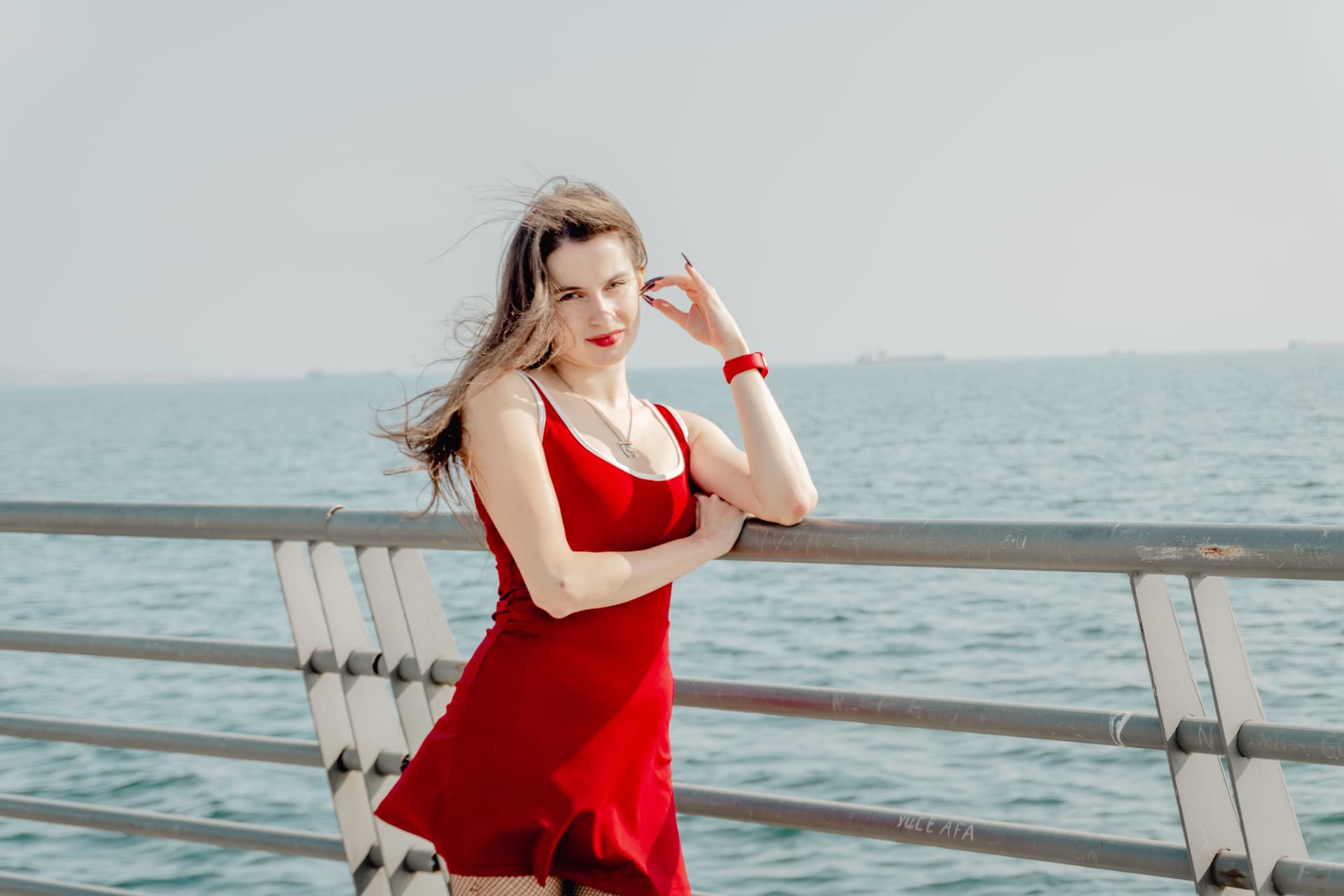 A woman in a red sundress with white trim leans against a railing on a sunlit pier, the open sea behind her.